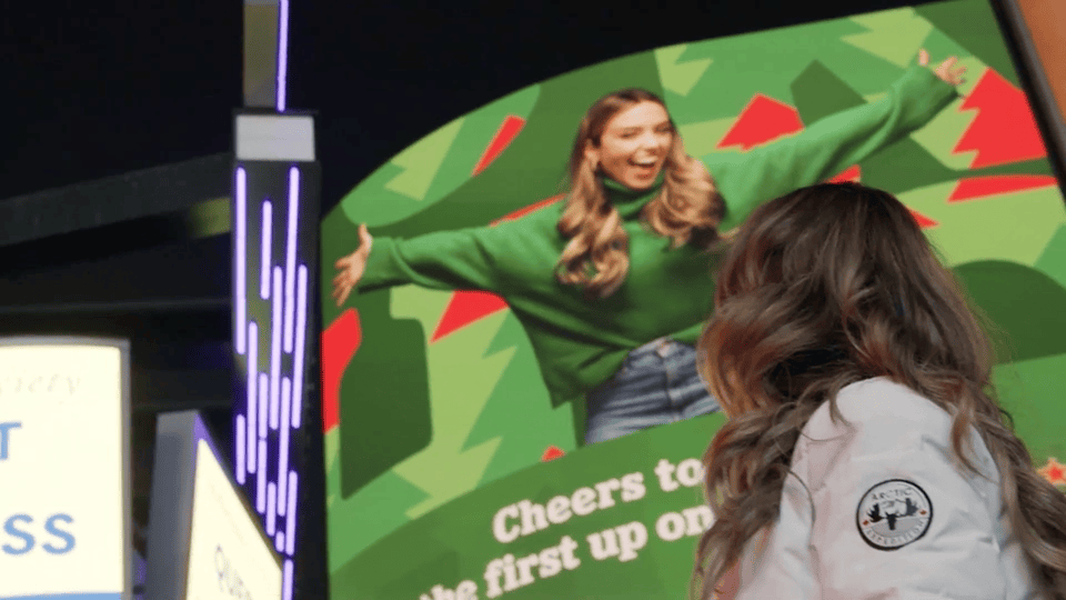 Animated gif of a blonde woman pointing over her shoulder to a Heinken holidays branded billboard with her image in Yonge Dundas square.