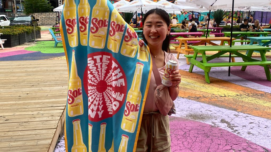 A woman holding a can of Sol beer and Sol banner at a sunny colourful patio.